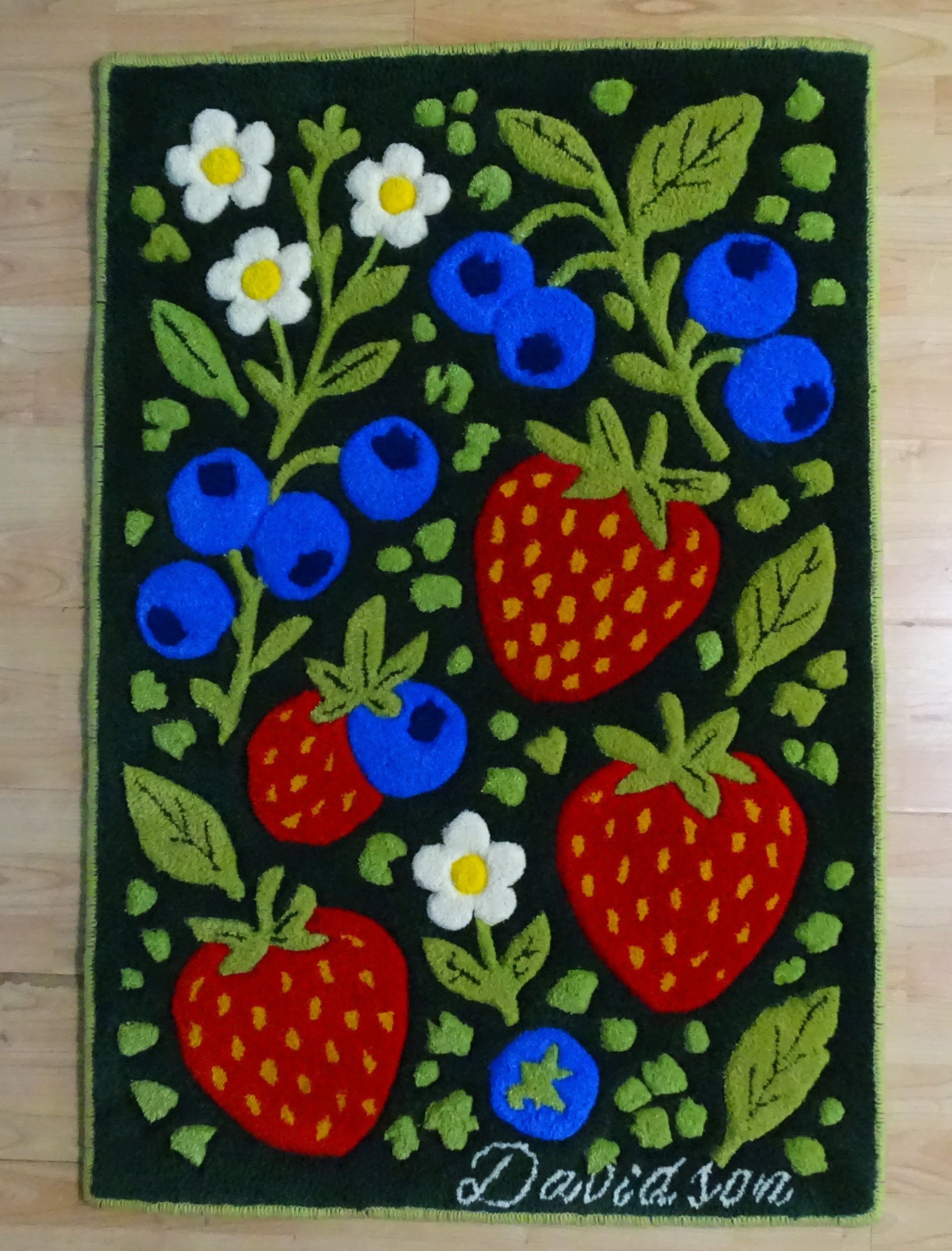 Handgefertigter personalisierter Wollteppich mit Erdbeeren, Blaubeeren und Blumen aus 100 % baltischer Schurwolle, mit Wunschname, auf Holzboden fotografiert