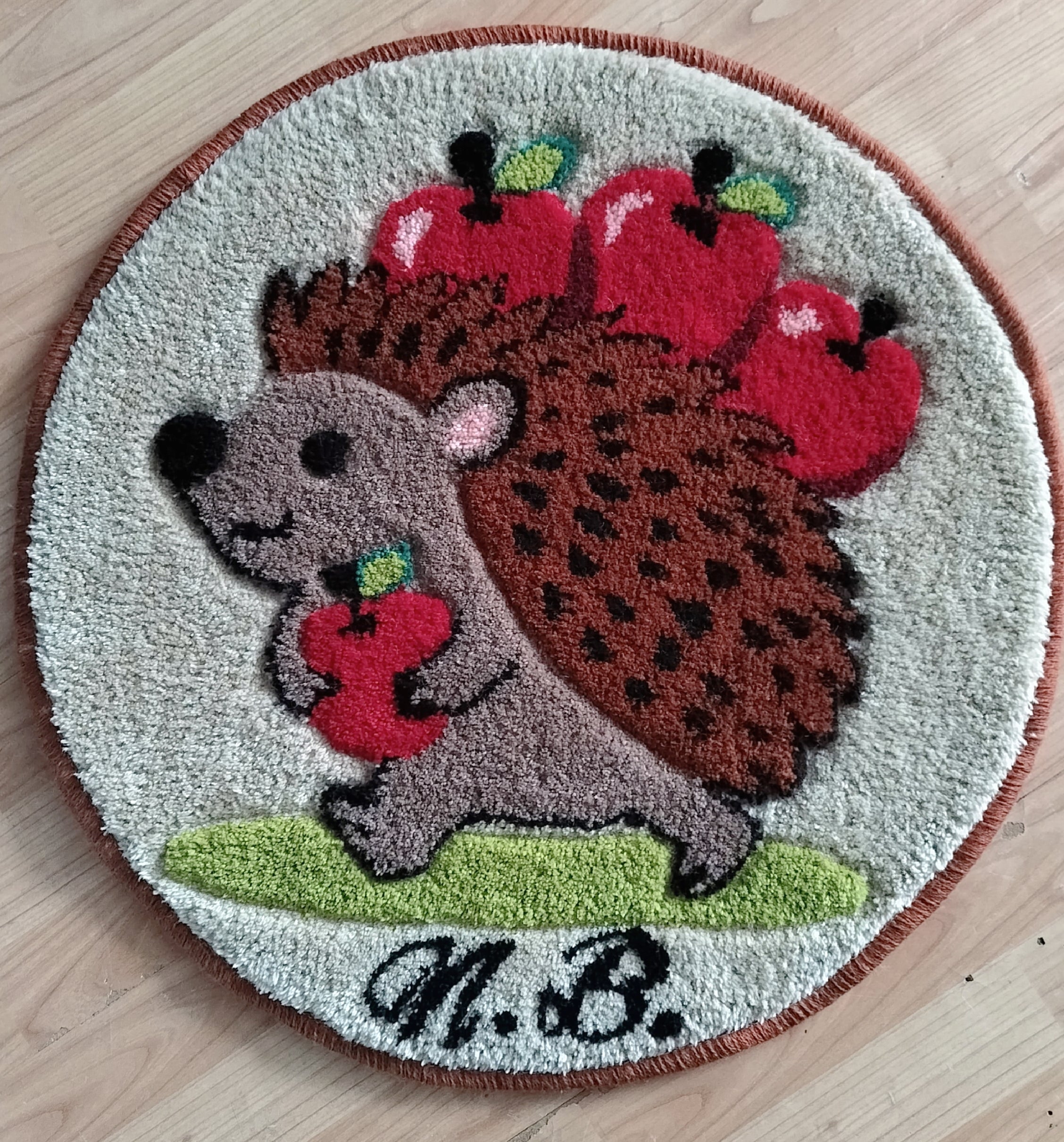 Runder handgetufteter Wollteppich mit Igel-Motiv und Äpfeln, personalisiert mit Initialen, auf Holzfußboden fotografiert.